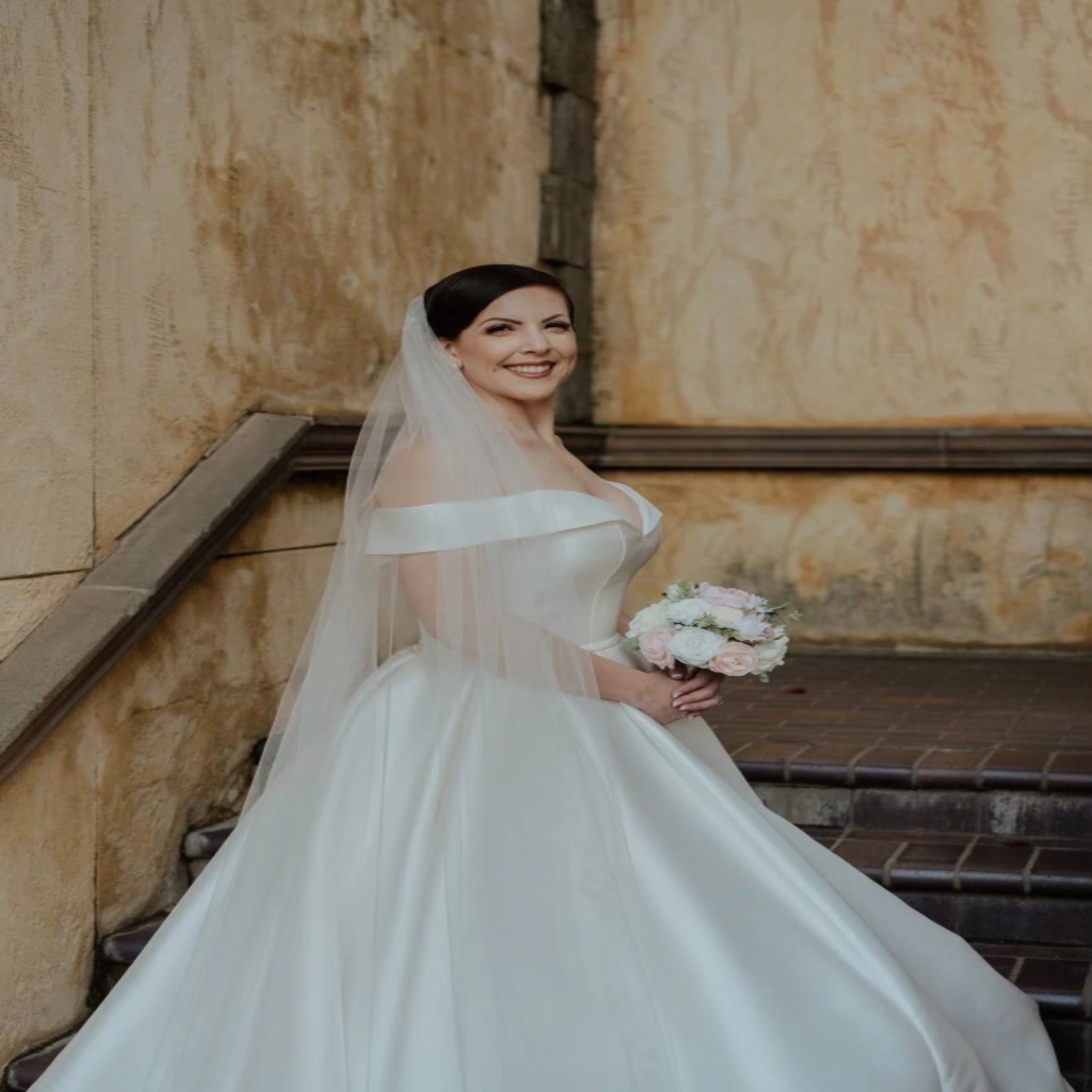 A bride in a full satin ballgown and cathedral veil on sandstone steps, holding a blush bouquet