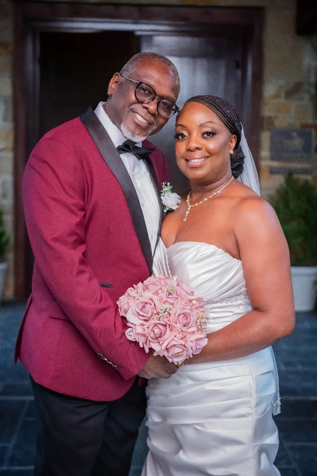 A bride and groom in a burgundy tuxedo with a blush rose bouquet, photographed in the doorway