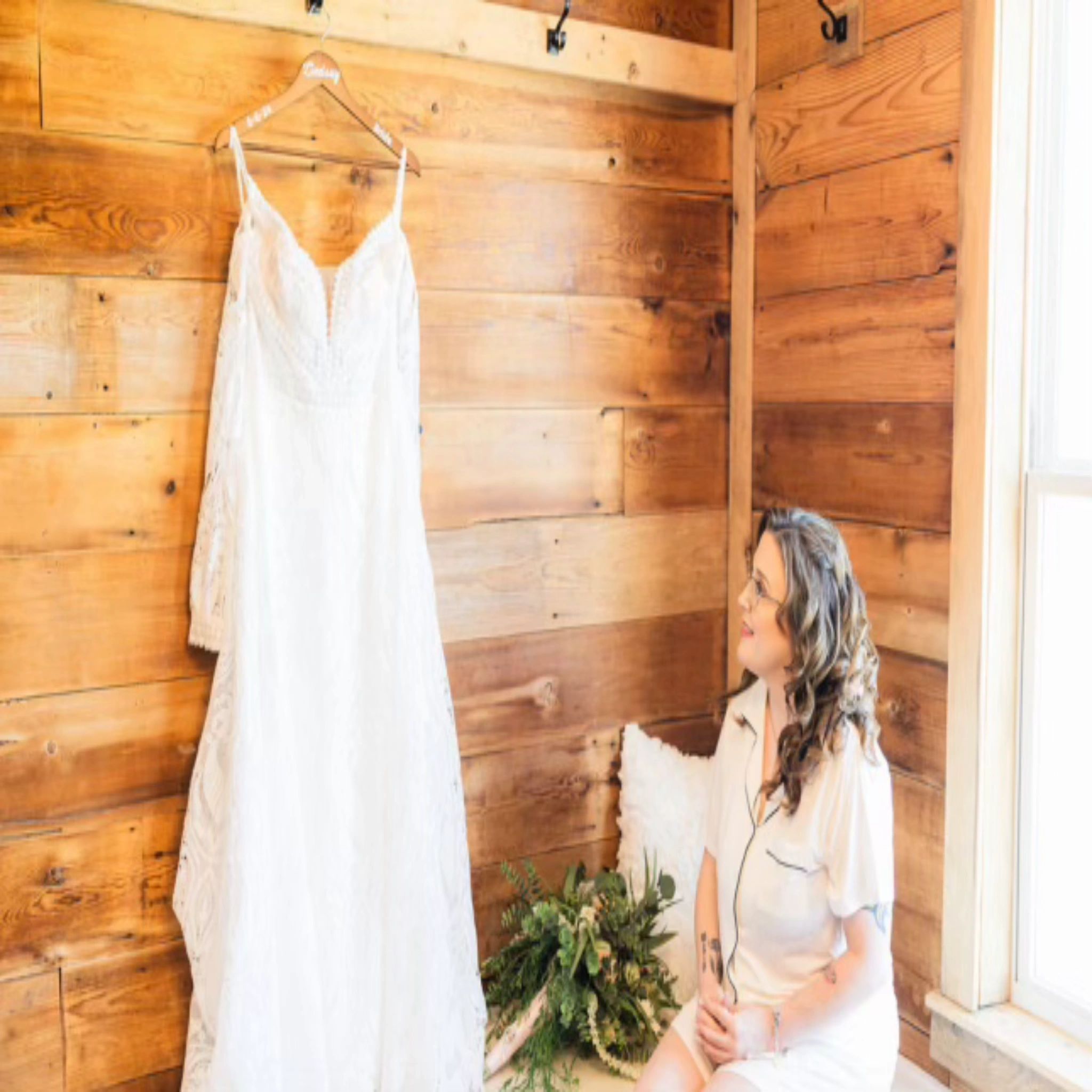 The wedding dress hanging on a timber wall while the bride sits beside it in the morning light